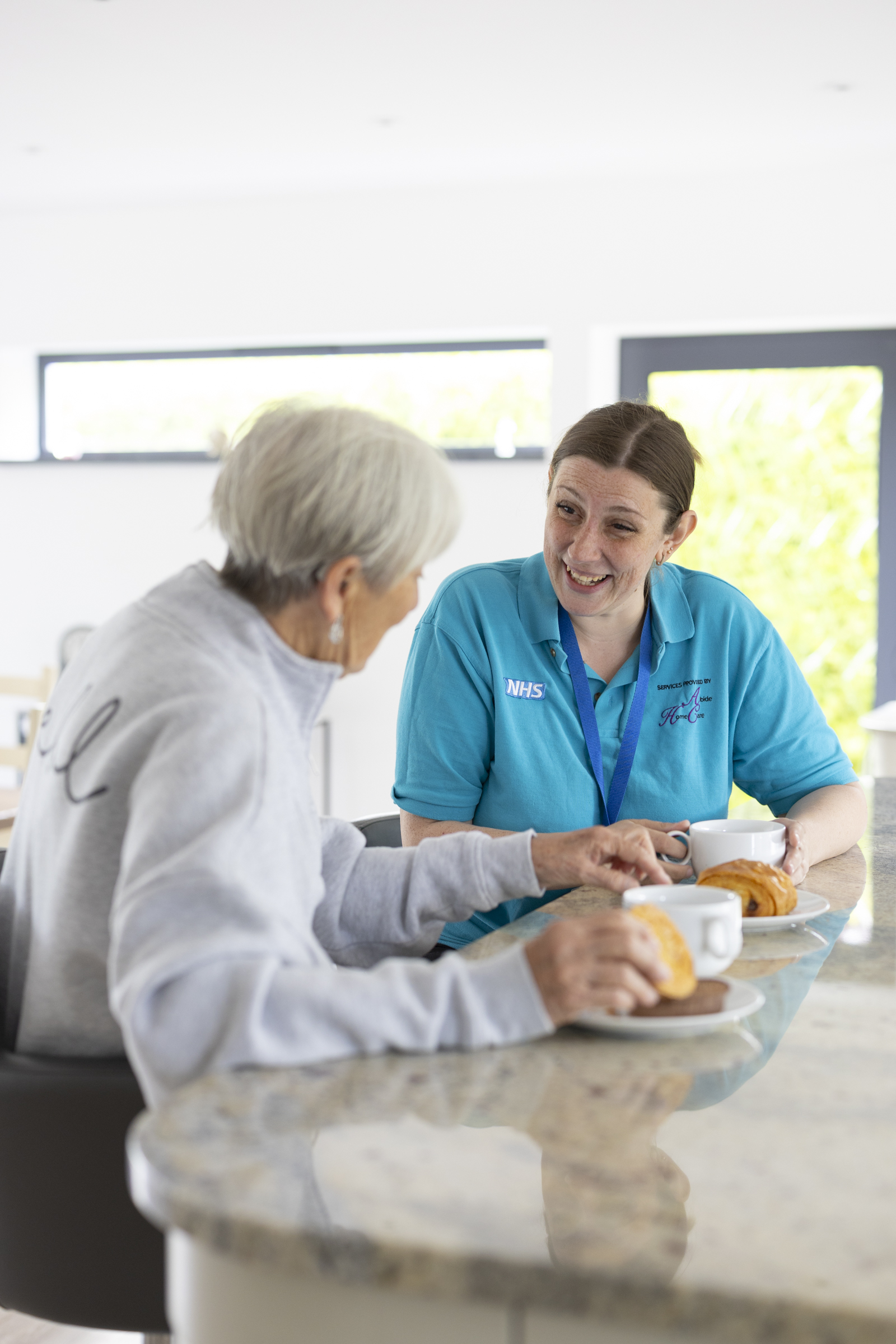 Abide care worker sharing tea and pastries while having a supportive conversation with an elderly woman at home.