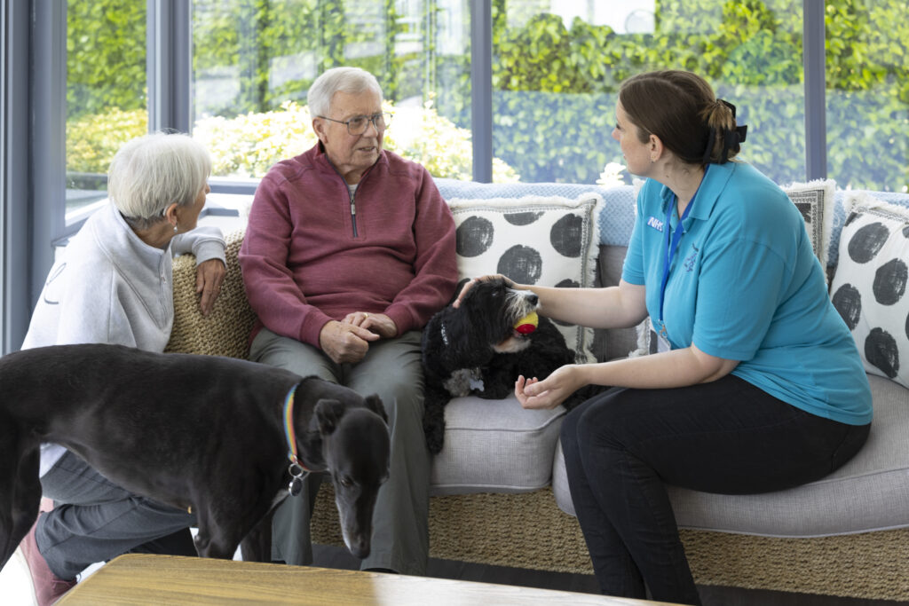 Caregiver chatting with an elderly couple and their pets in a bright home.