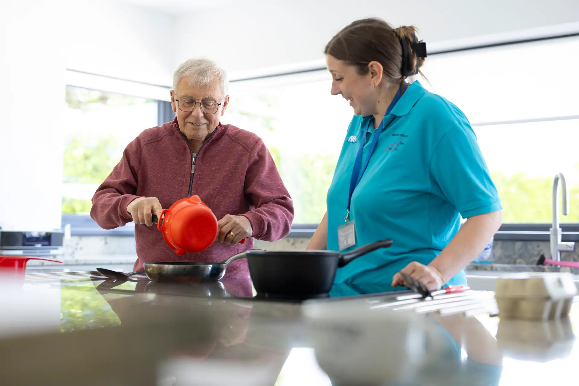 Care worker watching over an elderly man cooking in a kitchen.
