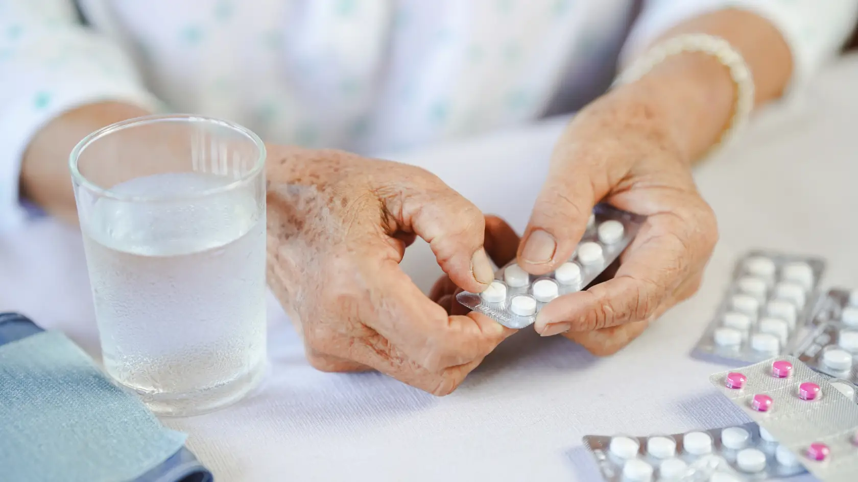 An elderly person's hands holding a blister pack of pills next to a glass of water.